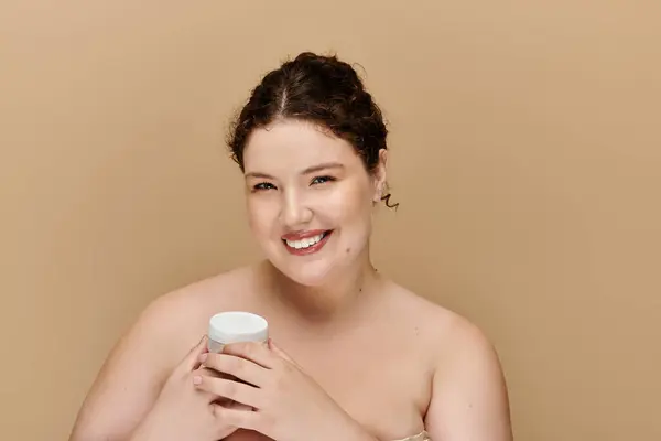 A beautiful plus size woman joyfully holds a cream jar, radiating confidence in soft beige light. — Stock Photo