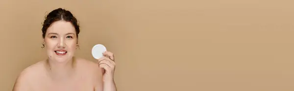 A beautiful plus size woman holds a skincare item, radiating confidence in a simple setting. — Stock Photo