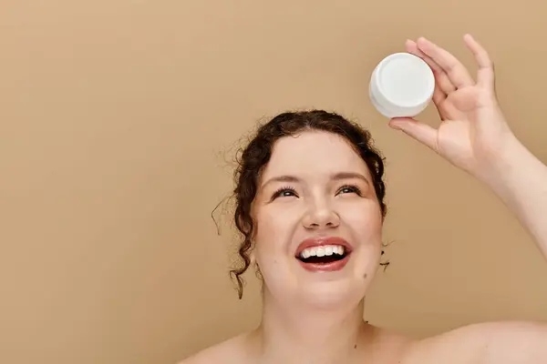 Una hermosa mujer de talla grande sonríe brillantemente mientras muestra un producto para el cuidado de la piel por encima de su cabeza. - foto de stock