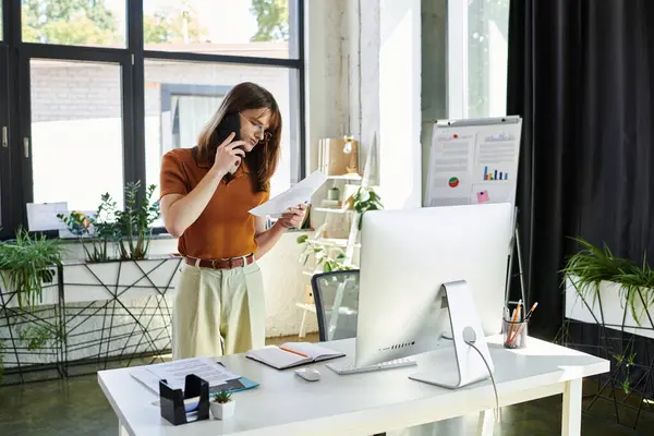 A young non binary person stands in a bright office, multitasking with documents and a phone. — Stock Photo