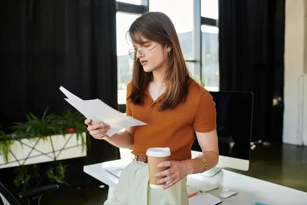 Una giovane persona non binaria si trova in un ufficio luminoso, leggendo note con una tazza di caffè in mano. — Foto stock