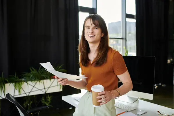 A young non binary individual stands in a stylish office, holding papers and a coffee cup. — Stock Photo