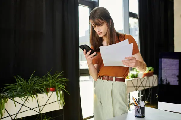 A focused individual reviews documents while using a smartphone in a contemporary workspace. — Stock Photo