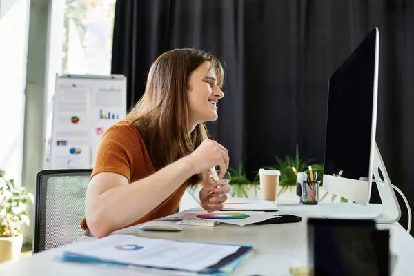 A non binary individual smiles while working on a project at their modern office desk. — Stock Photo
