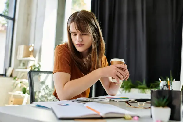 A young non binary person sits at a desk, sipping coffee in a vibrant workspace. — Stock Photo