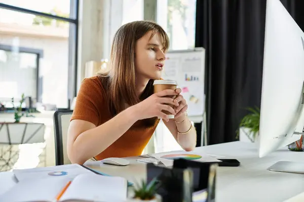 Concentrated young non binary person holds a coffee while engaging with a computer screen. — Stock Photo