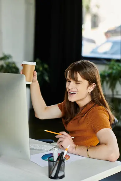 A young non binary individual smiles brightly while raising a coffee cup at their workspace. — Stock Photo