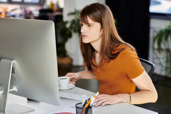 En una oficina moderna, una persona no binaria se involucra intensamente con su computadora mientras bebe café. - foto de stock
