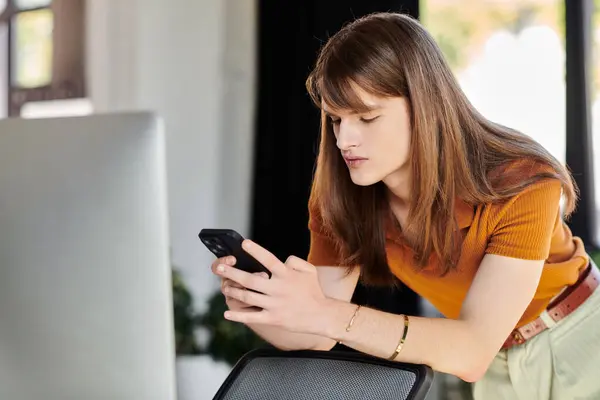 A young non binary individual checks their smartphone while leaning on an office desk. — Stock Photo