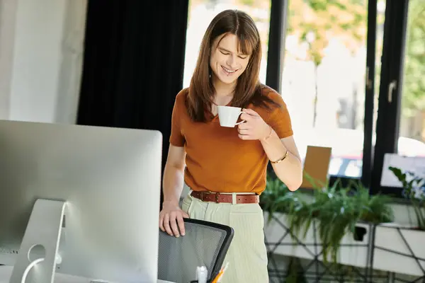 A non binary individual smiles while sipping coffee in a bright, stylish workspace. — Stock Photo
