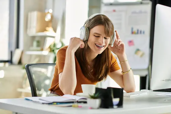 A young non binary person smiles joyfully with headphones on, immersed in their office work. — Stock Photo