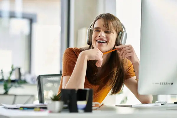 In a bright office, a young non binary individual smiles while working, wearing headphones. — Stock Photo