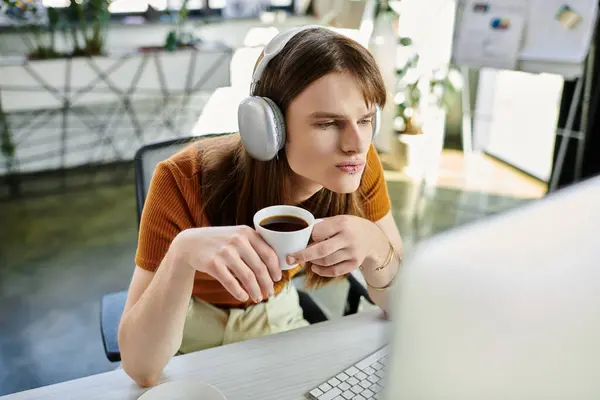 A young non binary person sips coffee and concentrates on their computer in a bright office. — Stock Photo