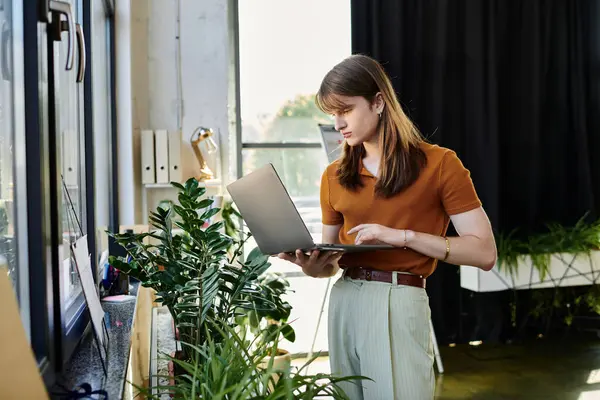 In a vibrant office space, a non binary person focuses intently on their laptop among lush plants. — Stock Photo