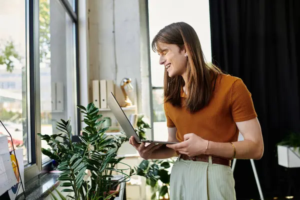 A young non binary person smiles while working on a laptop, surrounded by plants in a bright office. — Stock Photo