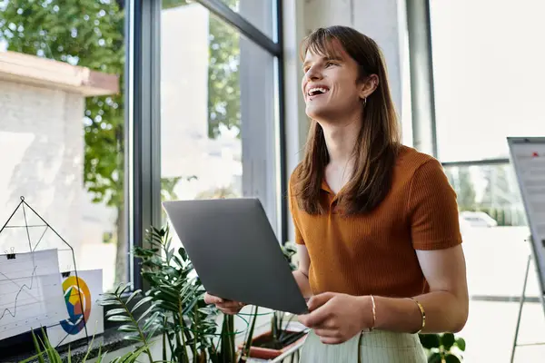 Une personne souriante non binaire se tient à un bureau, travaillant sur un ordinateur portable avec de la verdure autour d'elle. — Photo de stock