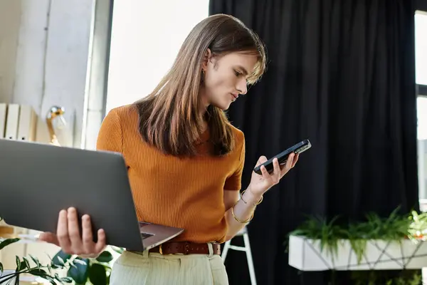 In un ufficio luminoso, una giovane persona non binaria destreggia tra laptop e telefono, focalizzata sul lavoro. — Foto stock
