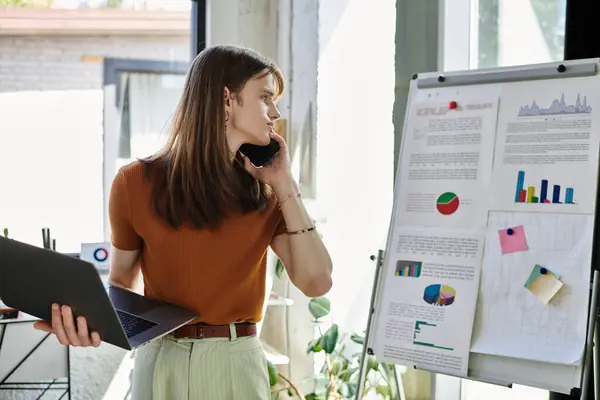 A young non binary person thoughtfully converses while holding a laptop and observing charts. — Stock Photo