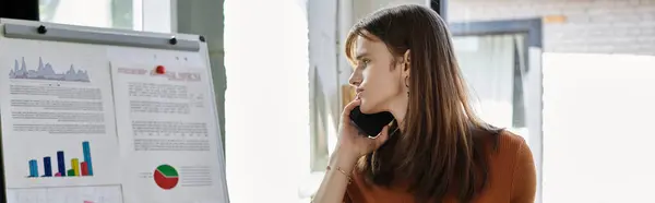Молодой не двоичный человек рассматривает отчеты и обсуждает детали работы по телефону. — стоковое фото