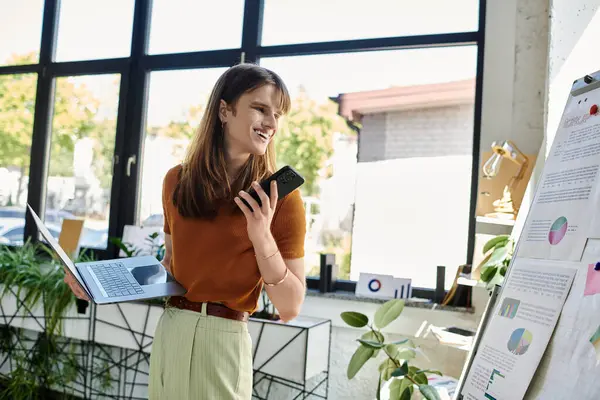 Un jeune individu non binaire sourit en multitâche avec un ordinateur portable et un smartphone dans un bureau. — Photo de stock