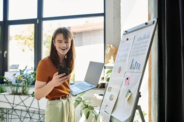 A young non binary person smiles while multitasking with a laptop and phone in a bright office. — Stock Photo