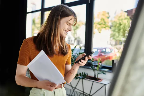 In an inviting office space, a young non binary person smiles at their phone while holding papers. — Stock Photo