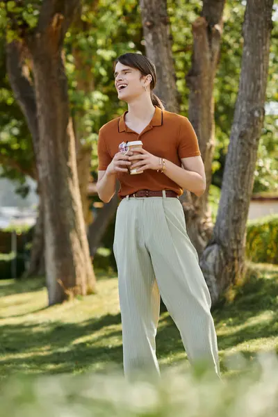 A young non binary individual stands under trees, sipping coffee and sharing a joyful moment. — Stock Photo