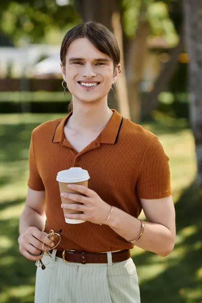 A young non binary person smiles while holding a coffee cup, surrounded by greenery. — Stock Photo