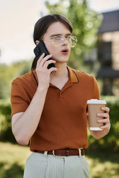 A young non binary individual talks on a smartphone, holding coffee outdoors in an office setting. — Stock Photo