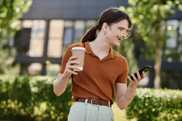 Un giovane individuo non binario sorride mentre controlla il telefono, la tazza di caffè in mano, all'aperto. — Foto stock