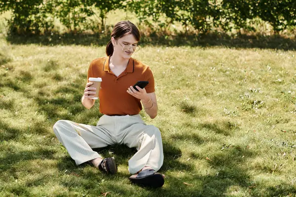 A non binary person sits on the grass, sipping coffee and using their phone. — Stock Photo