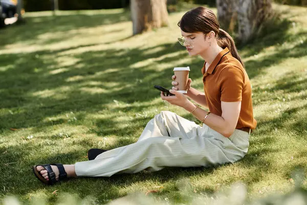 Sitting on the grass, a non binary individual sips coffee while engaging with a smartphone in hand. — Stock Photo