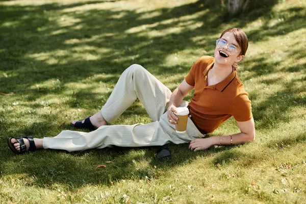 This young non binary individual sits on the grass, sipping coffee and smiling joyfully. — Stock Photo