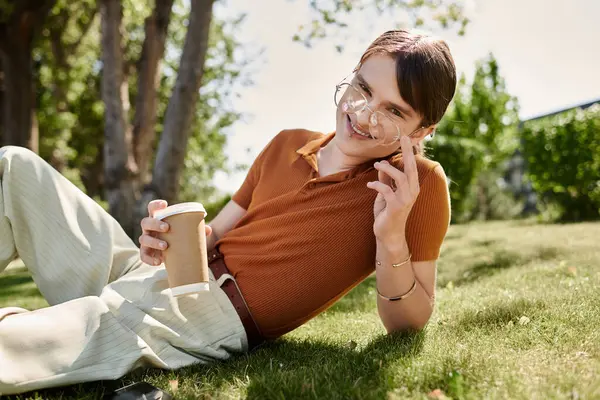 Relaxing outdoors, a non binary individual sips coffee while lounging in a cheerful office garden. — Stock Photo