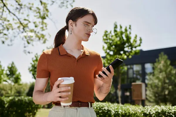 A young non binary individual holds a coffee cup and checks their phone, surrounded by greenery. — Stock Photo
