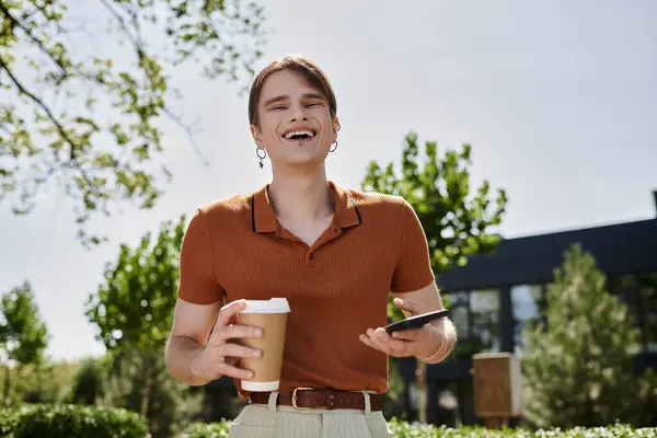 A cheerful non binary individual stands outdoors, holding a coffee and phone, enjoying the sun. — Stock Photo