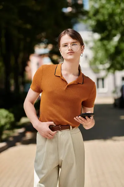 Eine junge, nicht binäre Person steht selbstbewusst im Freien und hält ein Smartphone im Büro. — Stockfoto