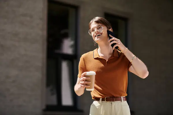 A young non binary person chats on the phone, holding a coffee cup in a stylish office environment. — Stock Photo