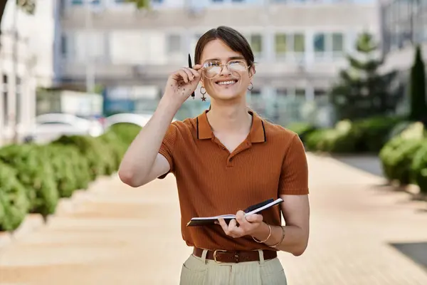 In an office park, a young non binary individual is cheerfully writing notes while smiling. — Stock Photo
