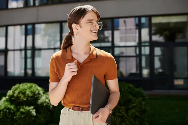 Spazio ufficio luminosamente illuminato ospita un individuo non binario in possesso di un computer portatile e sorridente gioiosamente. — Foto stock