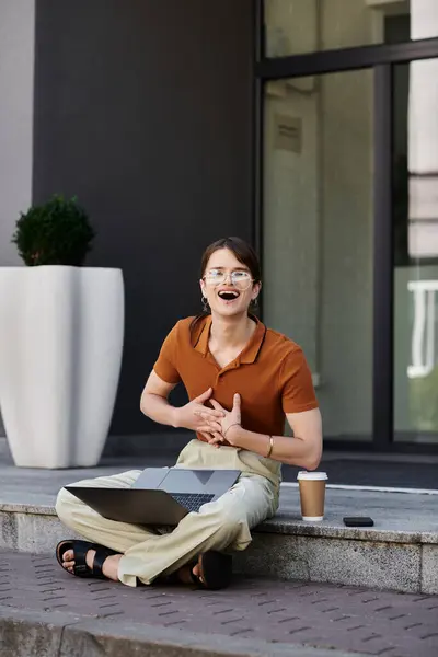 Una persona joven no binaria trabaja felizmente en su computadora portátil mientras toma café en los escalones. - foto de stock
