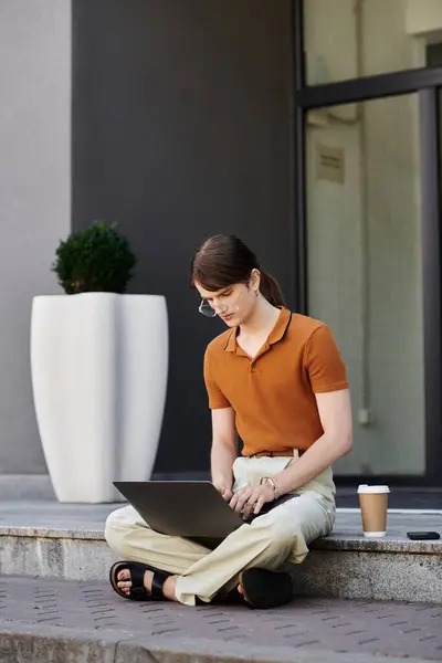 A young non binary individual sits on the steps, focused on their laptop, sipping coffee. — Stock Photo