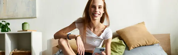 Une jeune femme dans la vingtaine sourit en s'asseyant sur son lit, s'engageant avec son téléphone dans une chambre confortable. — Photo de stock