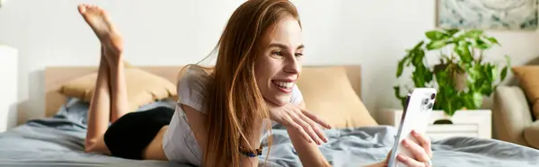 Una joven se involucra felizmente con su teléfono, tumbada en su cama en un espacio acogedor y luminoso, reflejando su vida cotidiana. — Stock Photo