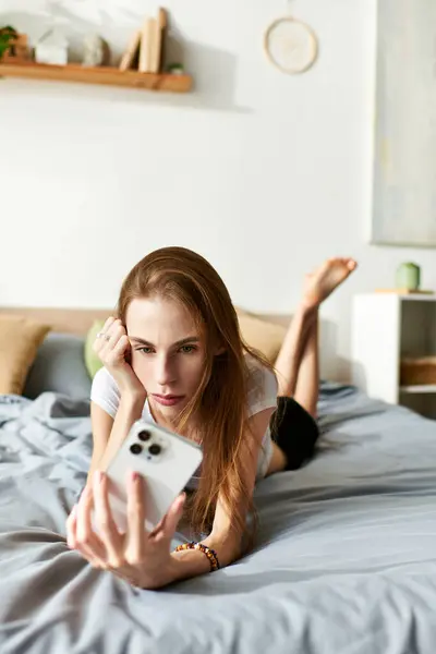 A young woman lies on her bed, absorbed in her thoughts while looking at her phone, contemplating her journey. — Stock Photo