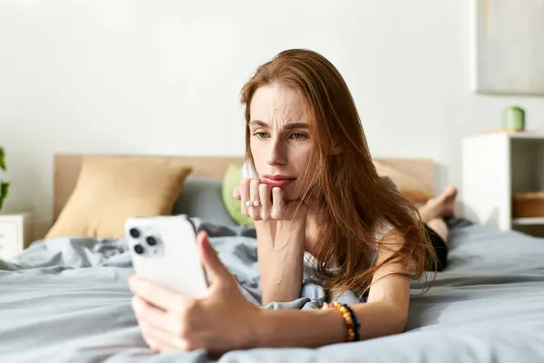 A young woman rests on her bed, deep in thought as she engages with her phone, navigating her struggles. — Stock Photo