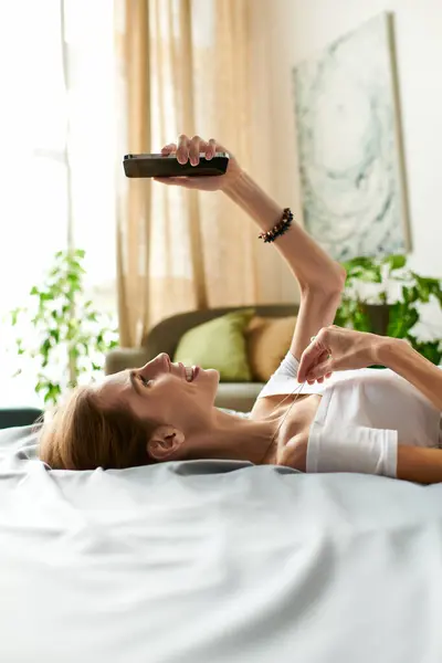 Une jeune femme se détend sur un lit, souriant alors qu'elle interagit avec son téléphone, embrassant un moment joyeux. — Photo de stock
