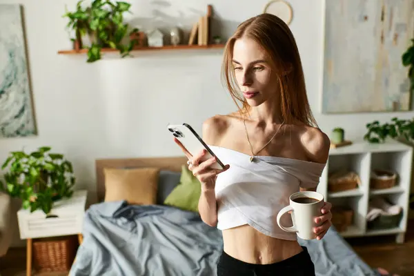 Молодая женщина с анорексией, потягивающая кофе, проверяя телефон посреди теплого, уютного пространства. — стоковое фото