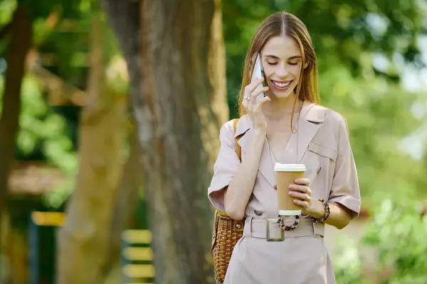 A young woman with a bright smile stands outdoors, chatting on her phone while savoring a coffee in hand. — Stock Photo