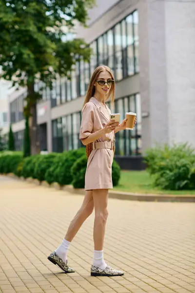 A young woman with an expressive smile strolls through an urban area, enjoying her coffee and the sunshine. — Stock Photo